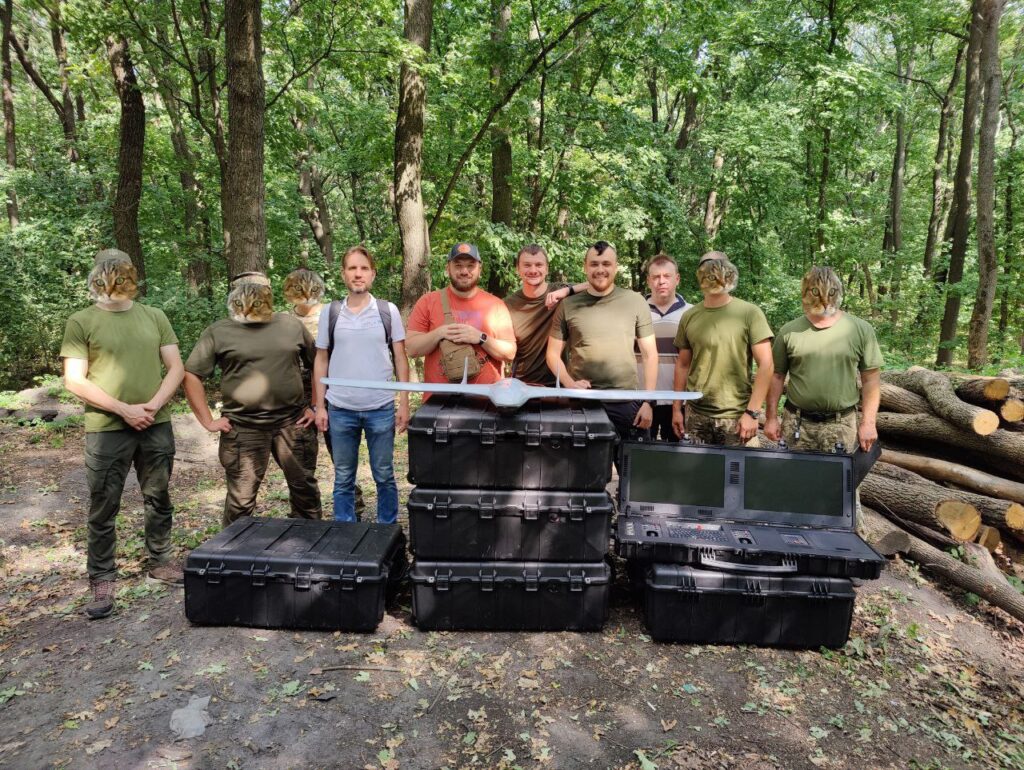 Команда на природі біля кейсів та безпілотника; обличчя учасників приховані.